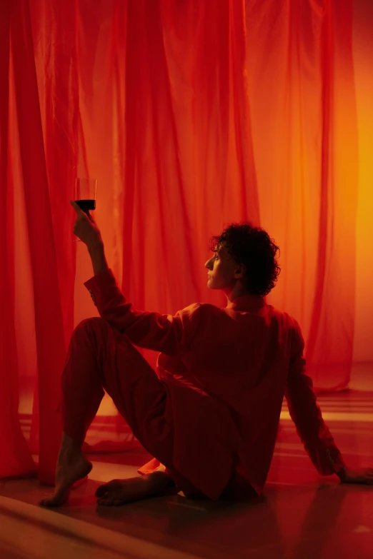 a woman sitting on the floor holding a glass of wine, inspired by Nan Goldin, magical realism, red and orange color scheme, finn wolfhard, glowing drapes, a portrait of issey miyake