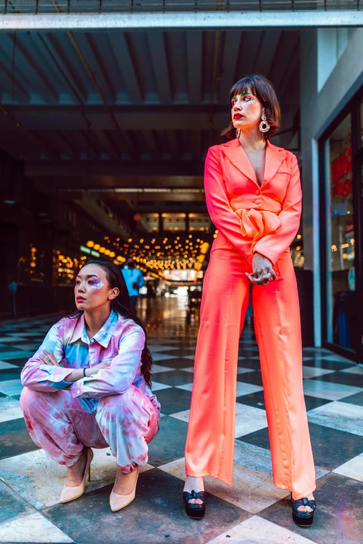 a couple of women standing next to each other on a tiled floor, an album cover, by Julia Pishtar, trending on pexels, neon colored suit, asian women, high rises, orange jumpsuit