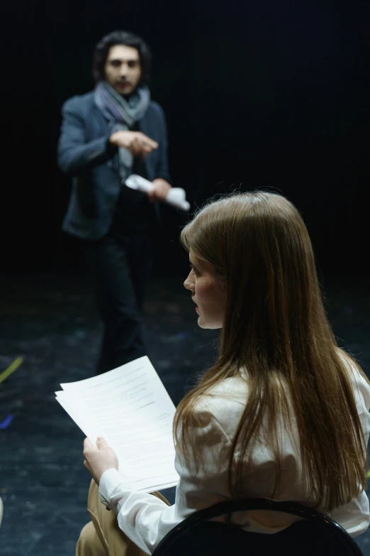 a woman sitting in a chair in front of a man, standing with a black background, document photo, performance, facing away