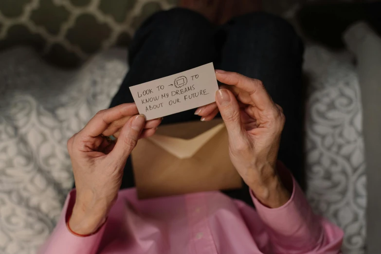 a woman sitting on top of a bed holding a piece of paper, by Julia Pishtar, pexels contest winner, private press, with some hand written letters, postapo game, holding gift, reassuring