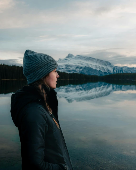 a woman standing in front of a lake with mountains in the background, by Emma Andijewska, he also wears a grey beanie, banff national park, photo of margot robbie, cold freezing nights