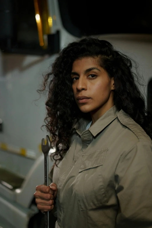 a woman standing in front of a truck holding a wrench, trending on pexels, serial art, imaan hammam, girl wearing uniform, serious expression, in a workshop