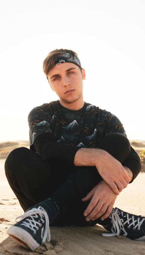 a man sitting on top of a sandy beach, inspired by Lucas Vorsterman, trending on pexels, graffiti, black beanie, patterned clothing, avatar image, good looking face