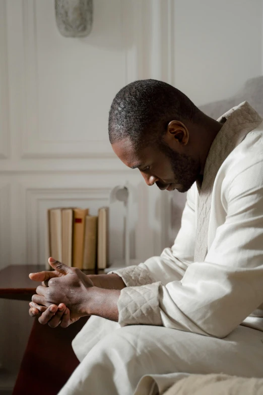 a man sitting on a couch praying, inspired by Theo Constanté, unsplash, idris elba as james bond, moroccan, wearing white pajamas, prayer hands