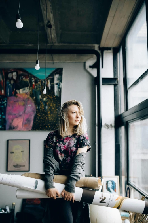 a woman holding a roll of paper in front of a window, a portrait, inspired by Louisa Matthíasdóttir, trending on unsplash, process art, young blonde woman, sitting on top a table, stood in a factory, prints