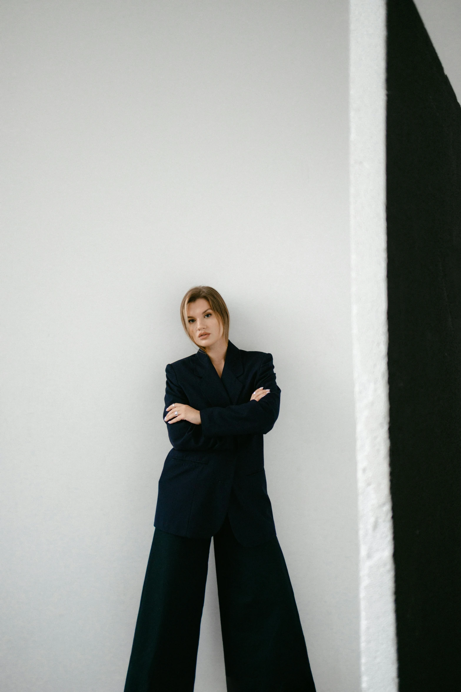 a woman leaning against a wall with her arms crossed, by Emma Andijewska, wearing causal black suits, kirsi salonen, plain background, asher duran