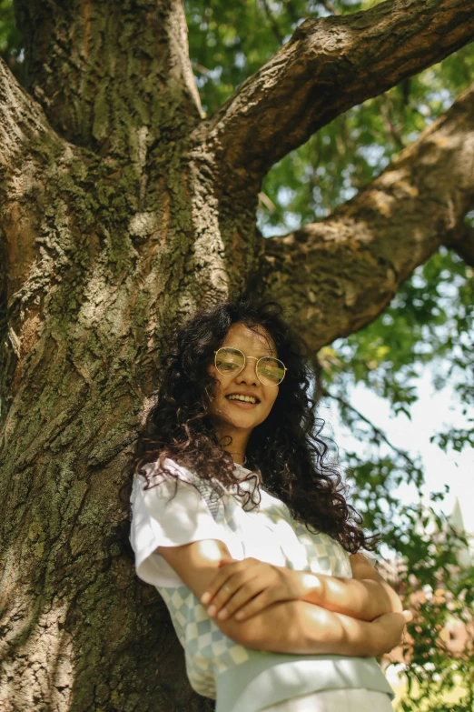 a woman leaning against a tree with her arms crossed, pexels contest winner, long wild black curly hair, ((trees)), girl wearing round glasses, an all white human