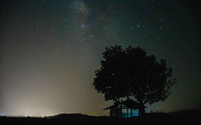 a tree sitting in the middle of a field under a night sky, pexels contest winner, light and space, wood cabin in distance, panorama view of the sky, stars and planets visible, distant mountains lights photo