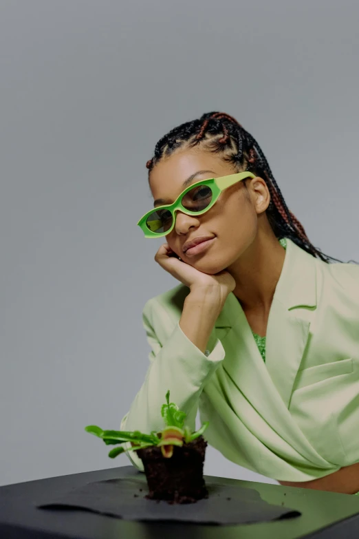 a woman sitting at a table with a plant in front of her, an album cover, inspired by Sophia Beale, trending on pexels, futuristic sunglasses, wearing green suit, cornrows, profile image