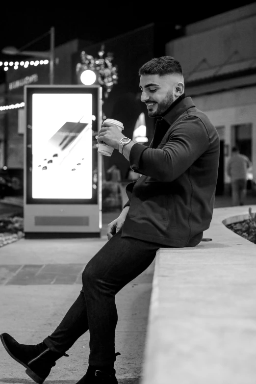 a man sitting on a ledge looking at his phone, a black and white photo, singer maluma, bus stop, holding a drink, slightly pixelated