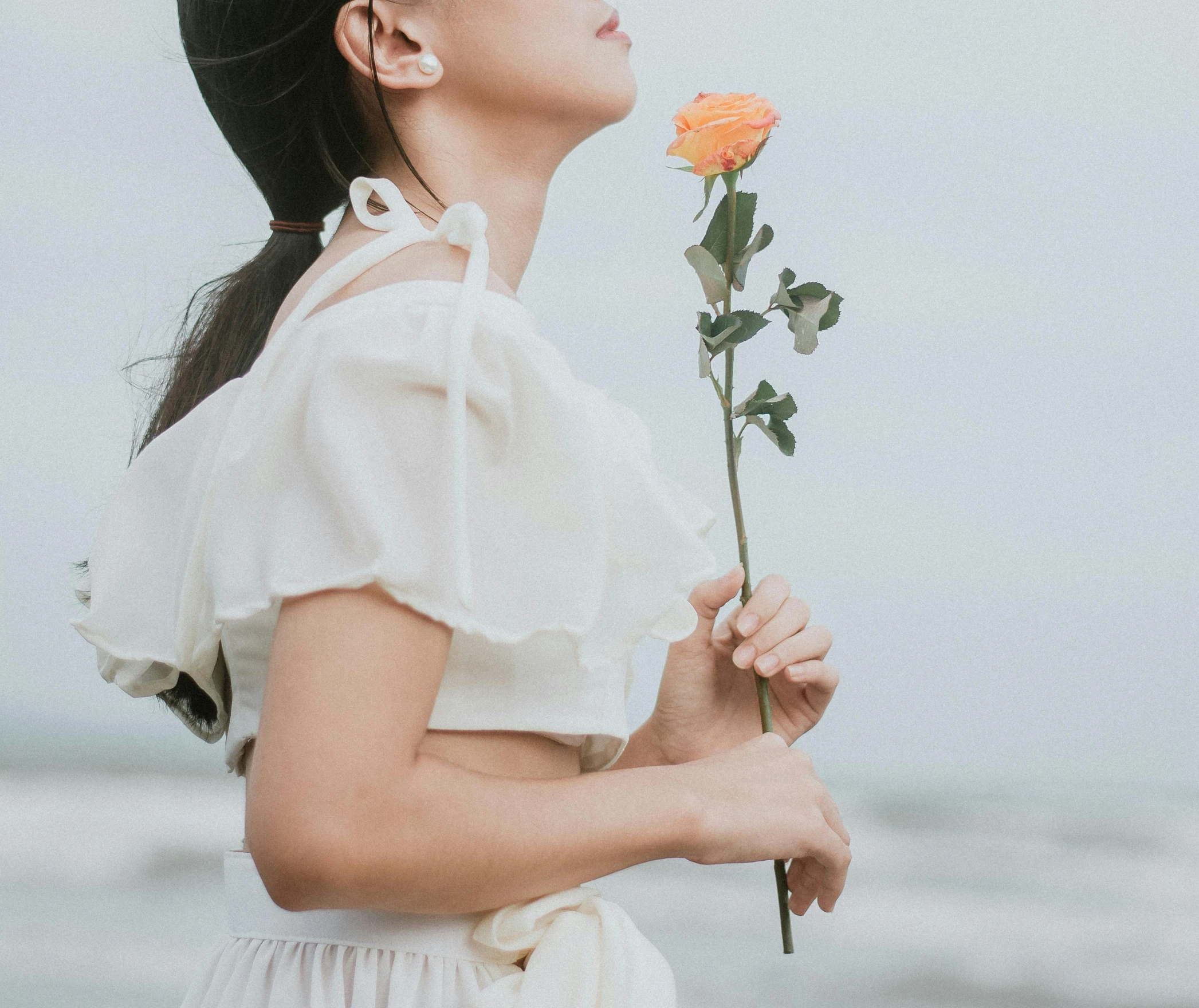 a woman standing on a beach holding a flower, inspired by Ruth Jên, unsplash, romanticism, white sleeves, young asian girl, holding a rose, white and orange