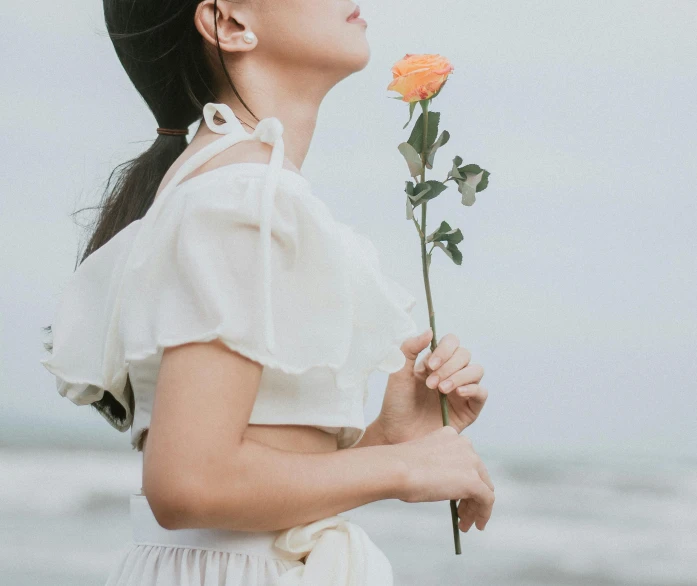 a woman standing on a beach holding a flower, inspired by Ruth Jên, unsplash, romanticism, white sleeves, young asian girl, holding a rose, white and orange