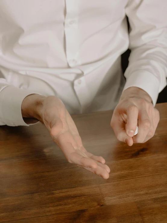 a man in a white shirt sitting at a table, by Adam Marczyński, offering the viewer a pill, reaching out to each other, natural wood top, using magic