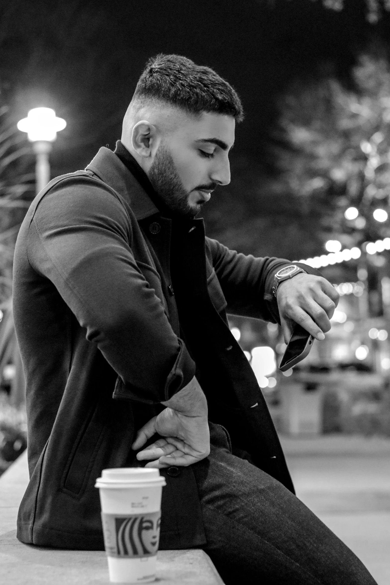 a man sitting on a bench looking at his cell phone, a black and white photo, hurufiyya, singer maluma, wearing a turtleneck and jacket, profile picture 1024px, man sitting facing away