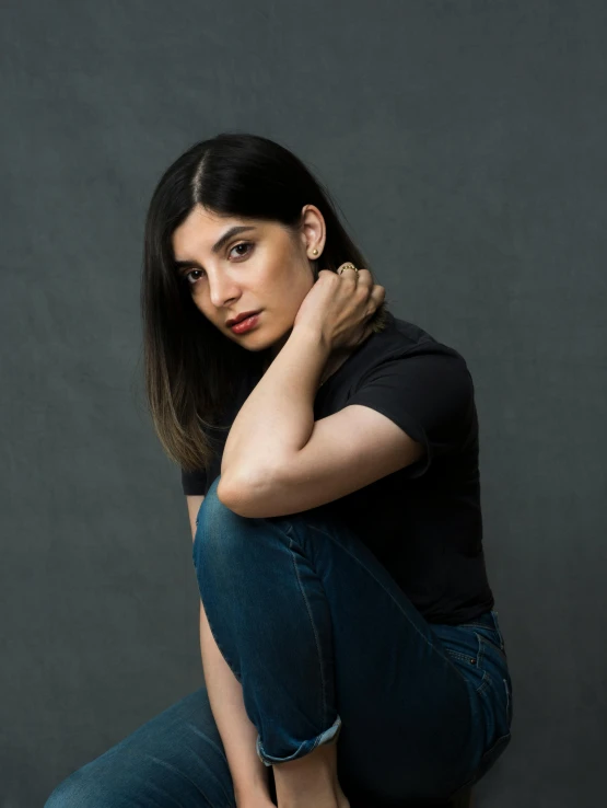 a woman sitting on a stool posing for a picture, an album cover, by Maryam Hashemi, wearing a dark shirt and jeans, dark. no text, headshot profile picture, christina kritkou