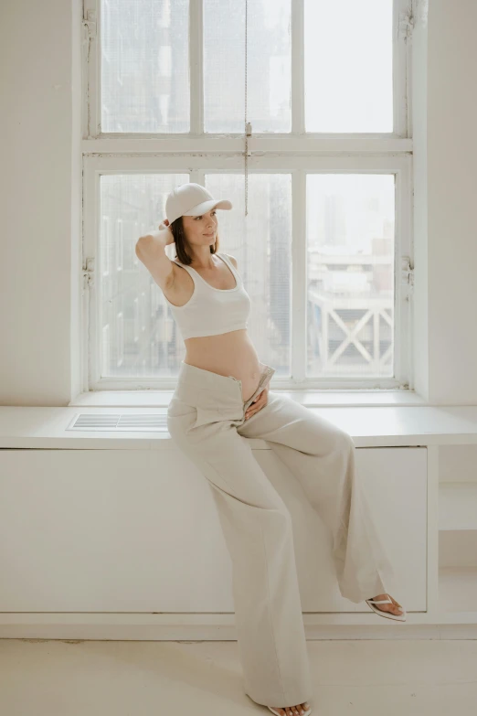 a pregnant woman sitting on a window sill, by Nicolette Macnamara, pexels contest winner, wearing a crop top, neutral background, white hat, baggy pants