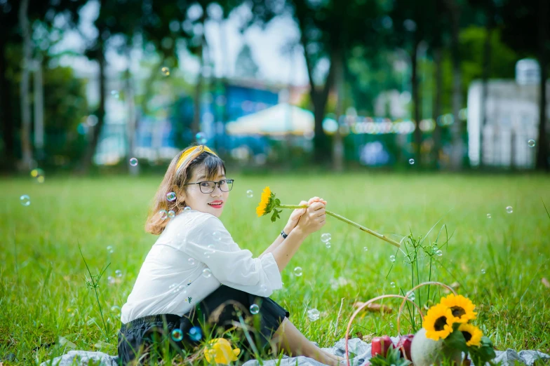 a woman sitting in the grass blowing bubbles, by Tan Ting-pho, unsplash, realism, wearing square glasses, with yellow flowers around it, avatar image, picnic