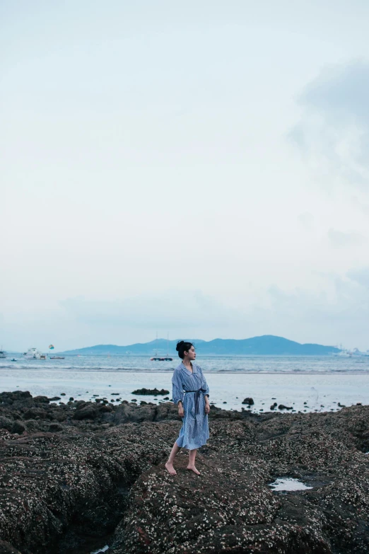 a woman standing on a rocky beach next to the ocean, a picture, inspired by Cui Bai, unsplash, bathrobe, in hong kong, blue toned, distant full body view
