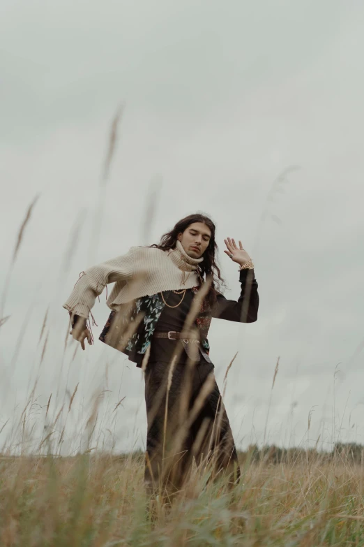 a woman standing in a field of tall grass, an album cover, trending on pexels, dressed with long fluent clothes, androgynous male, casting a protection spell, brown clothes