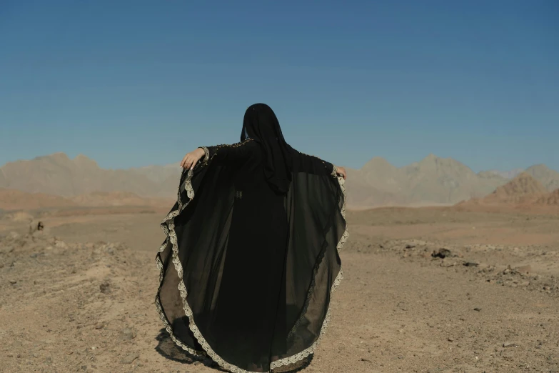 a person standing in the middle of a desert, an album cover, by Maryam Hashemi, symbolism, wearing black clothes and cape, still from a music video, rear-shot, ignant