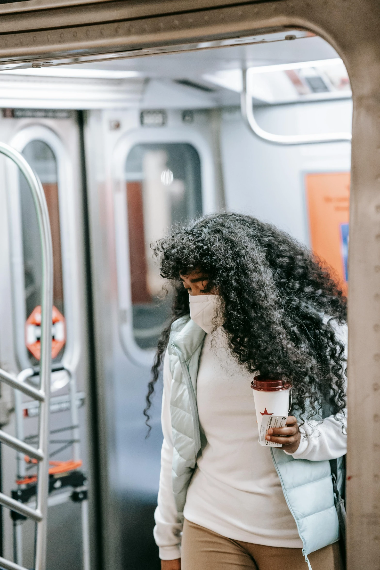 a woman standing next to a train holding a cup of coffee, by Carey Morris, pexels contest winner, black curly hair, face covered, nyc, sza