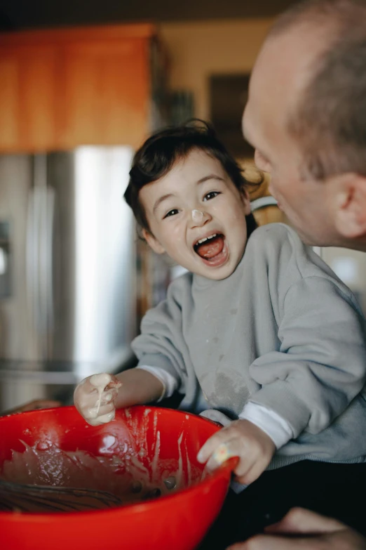 a man holding a child in a red bowl, pexels contest winner, head bent back in laughter, covered in white flour, cooking, thumbnail