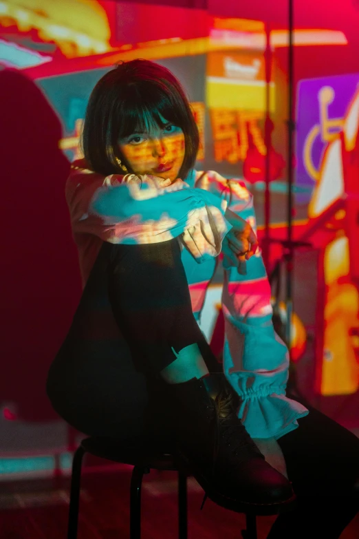 a woman sitting on a stool in front of neon signs, an album cover, inspired by Liam Wong, trending on pexels, holography, black haired girl wearing hoodie, performing on stage, kawaii playful pose of a dancer, vibrant scattered light