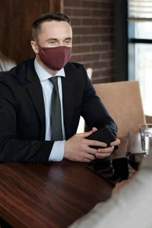 a man wearing a face mask sitting at a table, well dressed, premium quality, maroon, handheld