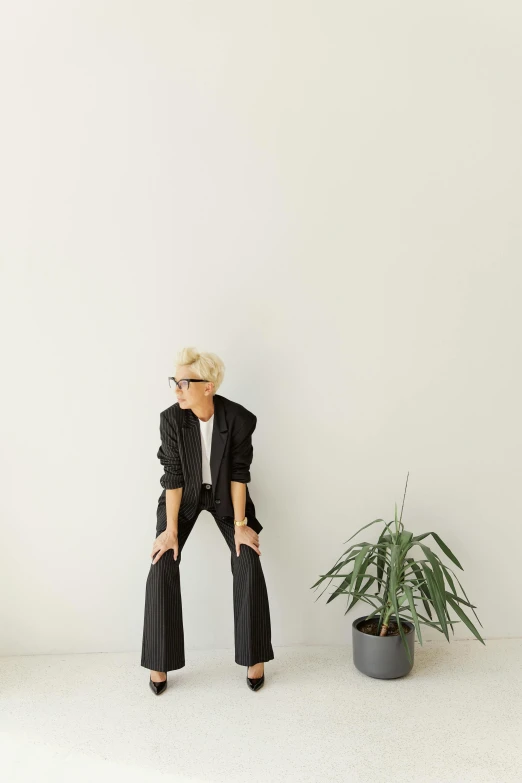 a woman leaning against a wall next to a potted plant, by Nina Hamnett, minimalism, in suit with black glasses, beth cavener, large pants, cai xukun