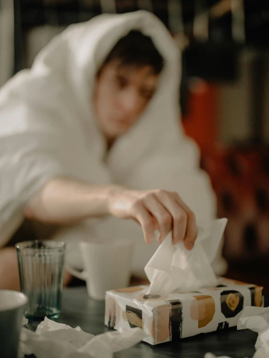 a woman sitting at a table in front of a box of tissues, by Julia Pishtar, pexels contest winner, happening, wearing white robe, gif, asian male, covered with blanket