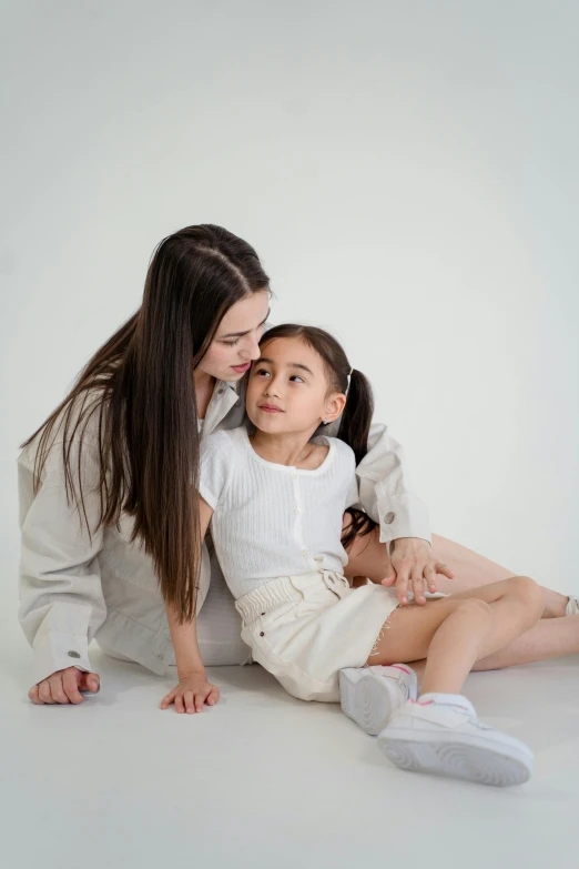 a woman sitting next to a little girl on the floor, light grey backdrop, holding each other, modelling, gen z