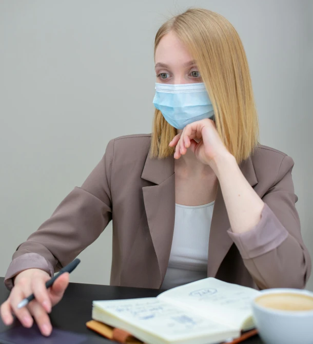 a woman wearing a face mask sitting at a table with a cup of coffee, pexels, office clothes, blonde women, russian academic, pastel'