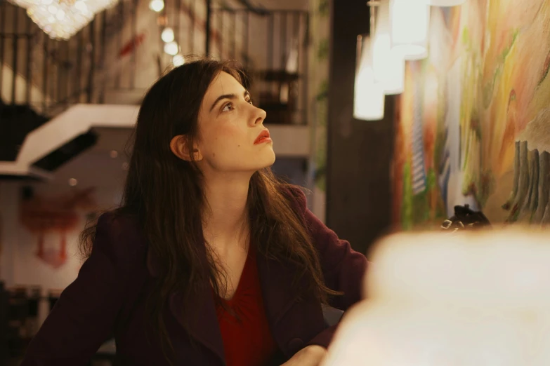 a woman sitting at a table in front of a painting, inspired by Elsa Bleda, pexels contest winner, serial art, portrait of alexandra daddario, city lights, looking to his side, as she looks up at the ceiling