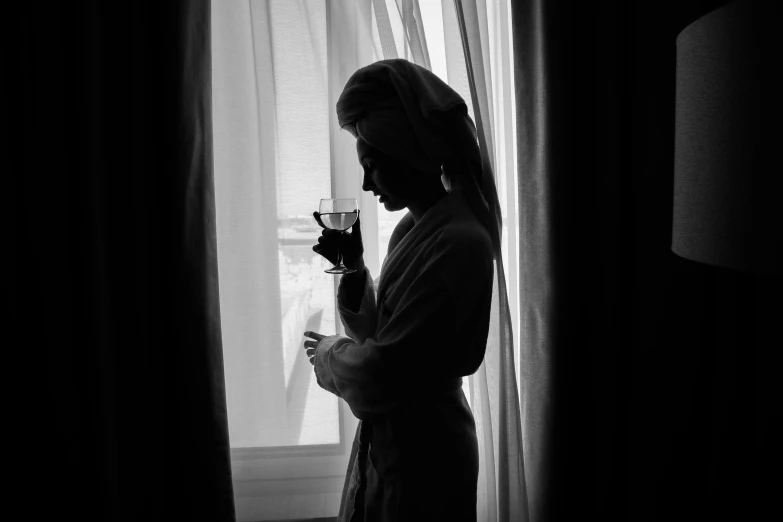 a woman standing in front of a window holding a glass, a black and white photo, by John Hutton, pexels, wearing a robe, character silhouette, maxim sukharev, hotel room