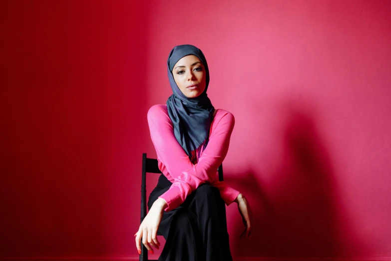 a woman in a hijab sitting on a chair, inspired by Maryam Hashemi, pexels contest winner, hurufiyya, hot pink and black, casual pose, in red background, wearing casual clothing