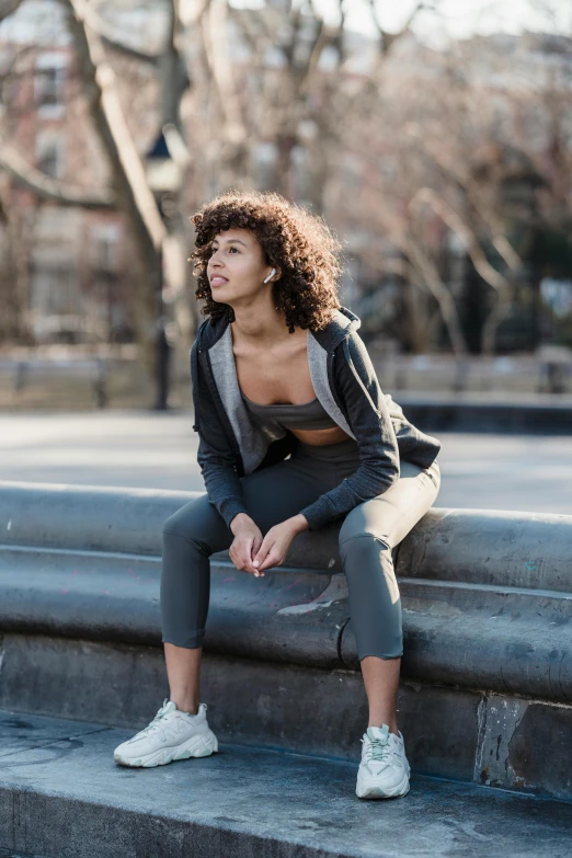 a woman sitting on a ledge in a park, a portrait, by Leo Michelson, trending on unsplash, happening, two piece workout clothes, (dark shorter curly hair), grey clothes, ny