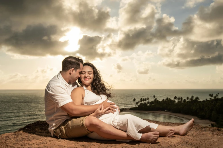 a man and woman sitting on top of a hill next to the ocean, a portrait, by Dan Luvisi, pexels contest winner, pregnancy, puerto rico, avatar image, warm glow