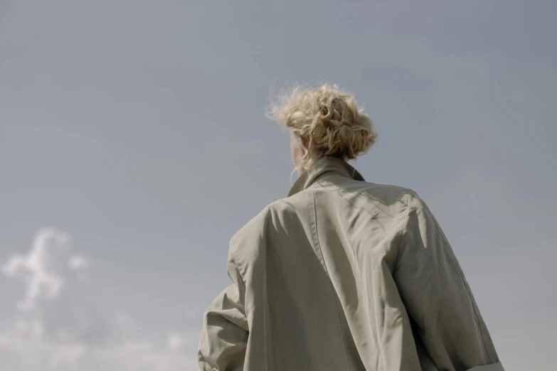 a man standing on top of a lush green field, an album cover, by Caro Niederer, unsplash, visual art, wearing a long beige trench coat, curly blond, low - angle shot from behind, grey sky