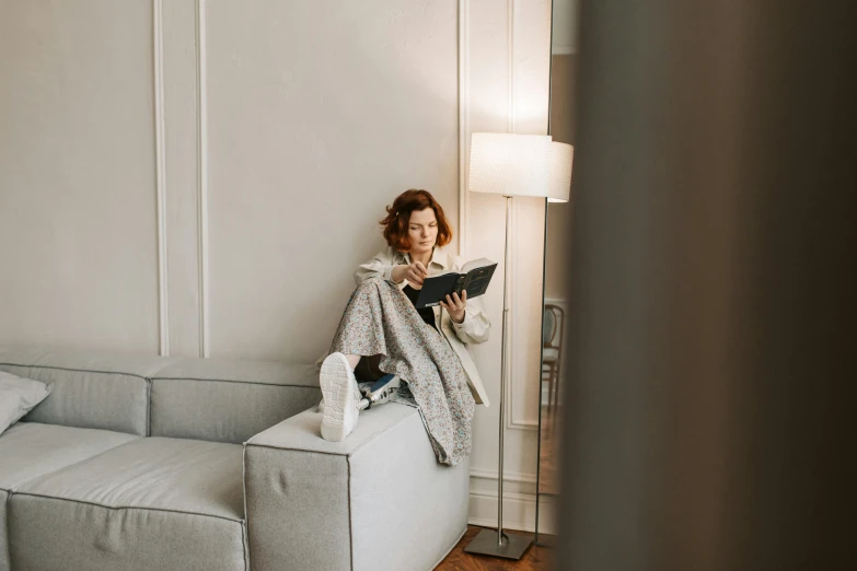 a woman sitting on a couch reading a book, by Emma Andijewska, grey robe, high quality upload, at the sitting couch, european woman photograph