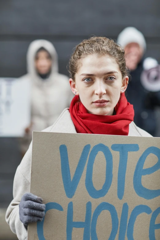 a woman holding a sign that says vote choice, a photo, by Harriet Zeitlin, shutterstock, renaissance, thin young male, lorde, mittens, looking straight to camera