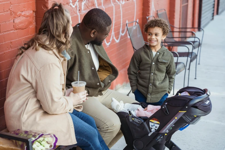 a woman sitting on a bench next to a child in a stroller, by Everett Warner, pexels, portrait of family of three, in a sidewalk cafe, black man, promotional image