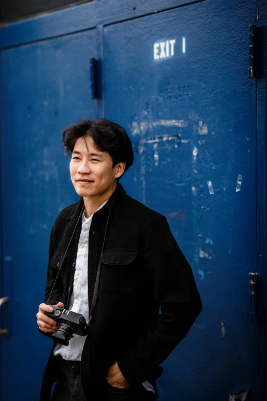 a man standing in front of a blue door holding a camera, inspired by Joong Keun Lee, halfbody headshot, standing in a city center, with a black background, sholim
