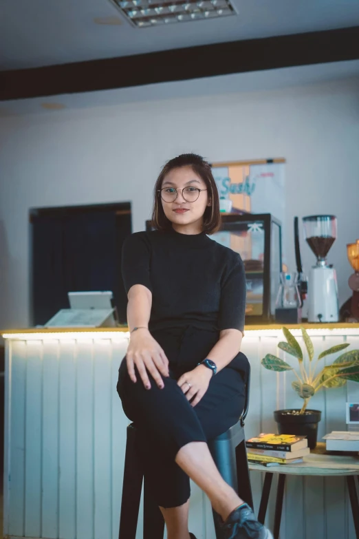 a woman sitting on a stool in a kitchen, a portrait, inspired by Ruth Jên, pexels contest winner, wearing a black shirt, in a coffee shop, wearing square glasses, malaysian