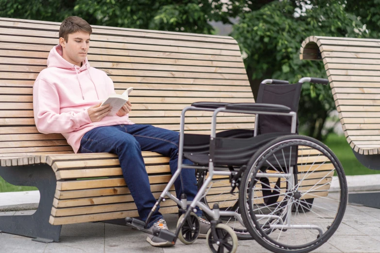 a man in a pink hoodie is sitting on a bench with a wheelchair, reading a book, enhanced quality, multi - coloured, bench