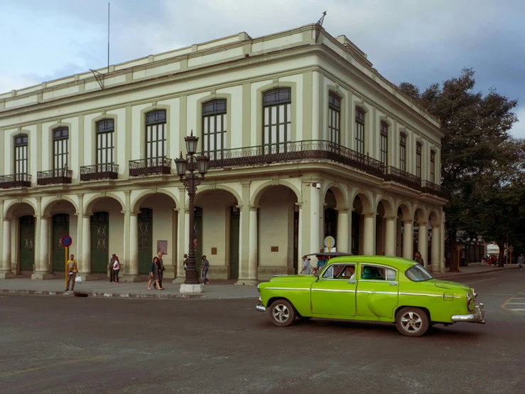 a green car driving down a street next to a tall building, inspired by Ceferí Olivé, neoclassicism, royal palace, tula, exterior view, square