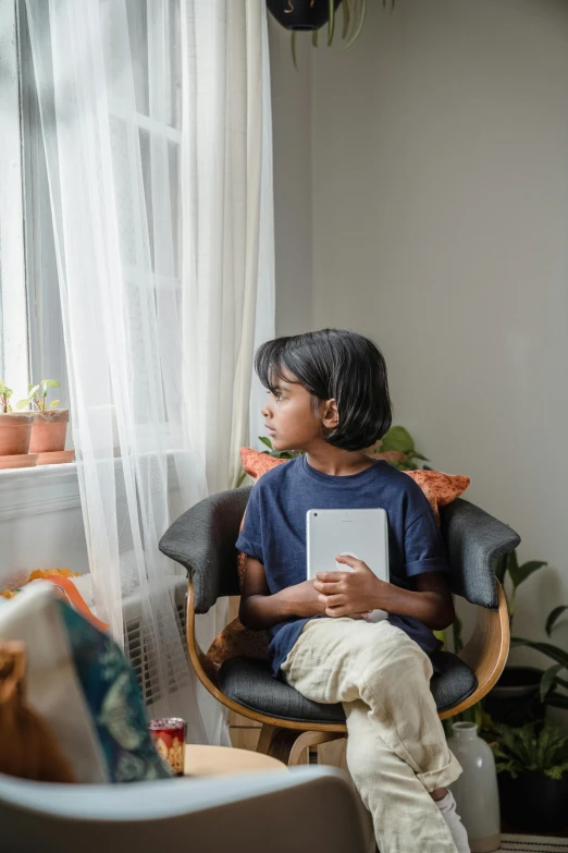 a little boy sitting in a chair holding a book, pexels contest winner, looking out window, technologies, young asian girl, in a living room