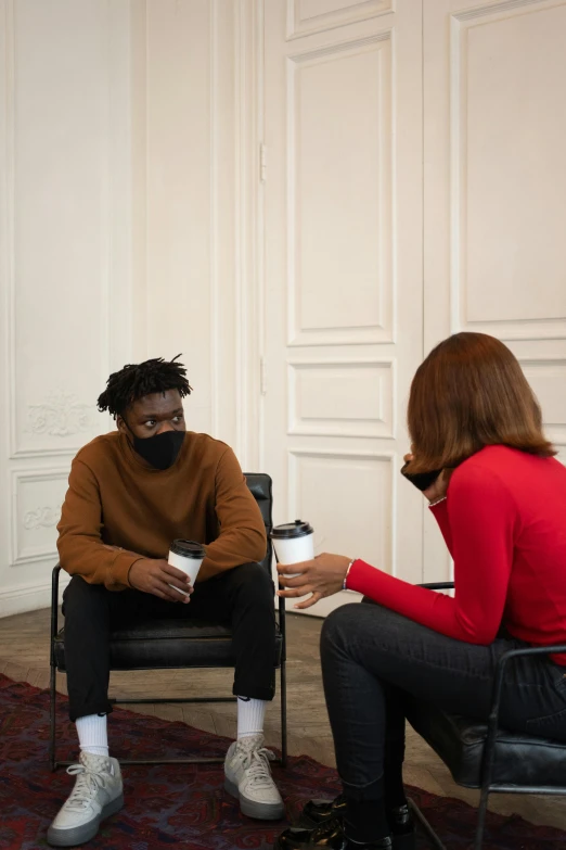 a group of people sitting in chairs in a room, two people, black man, drinking coffee, masked person in corner