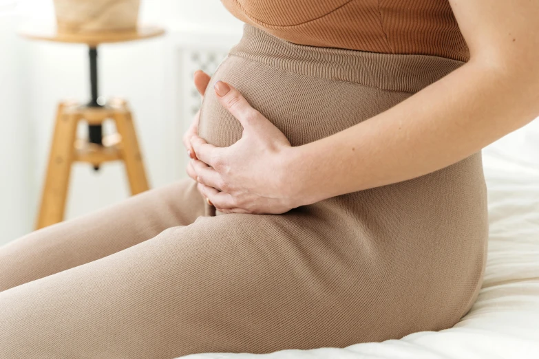 a woman sitting on a bed holding her stomach, trending on pexels, background image, manuka, bump in form of hand, brown