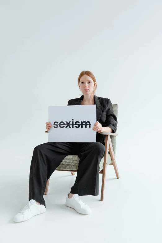 a woman sitting in a chair holding a sign that says sexism, trending on pexels, excessivism, on a gray background, with pale skin, redhead woman, profile image