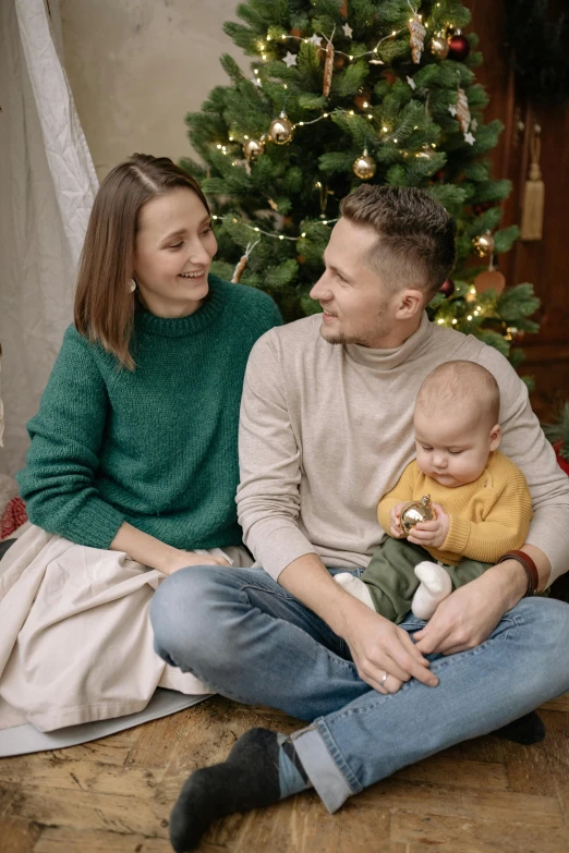 a man and woman sitting next to a baby in front of a christmas tree, by Adam Marczyński, pexels, renaissance, green and brown clothes, very handsome, gif, indoor scene